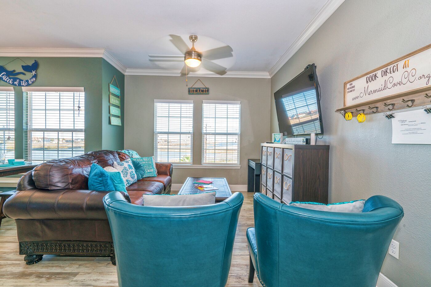 Een woonkamer met een bruine leren bank, teal fauteuils, een plafondventilator en een televisie op een houten dressoir.