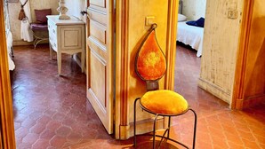 Una habitación con puerta de madera, una mesita de noche con lámpara, una silla con cojín y una cama con almohada azul.