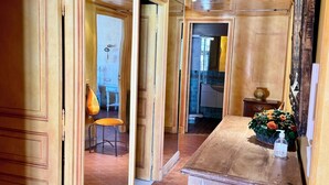 Un pasillo con puertas de madera y suelo de baldosas, que conduce a una habitación con una silla y una pequeña mesa con un jarrón y flores.