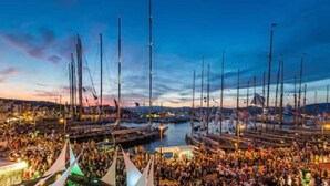 Un puerto deportivo con varias embarcaciones amarradas, una multitud de gente y un atardecer en el fondo.
