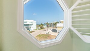 A view through a hexagonal window with a white frame, overlooking a residential area with houses and palm trees. - Turquoise Treasure - Boardwalk to the Beach! Beach Gear Credit I (Port Aransas)