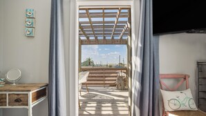 Ein Zimmer mit einem Holztisch, einem Stuhl, einem kleinen Tisch mit Spiegel, einem Korbstuhl und Blick auf eine Holzterrasse sowie Palmen draußen.