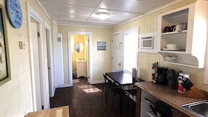 A narrow hallway leading to a bathroom with a sink and mirror. A dining table with chairs is in the foreground, and a kitchen area with a microwave and shelves is visible to the right.