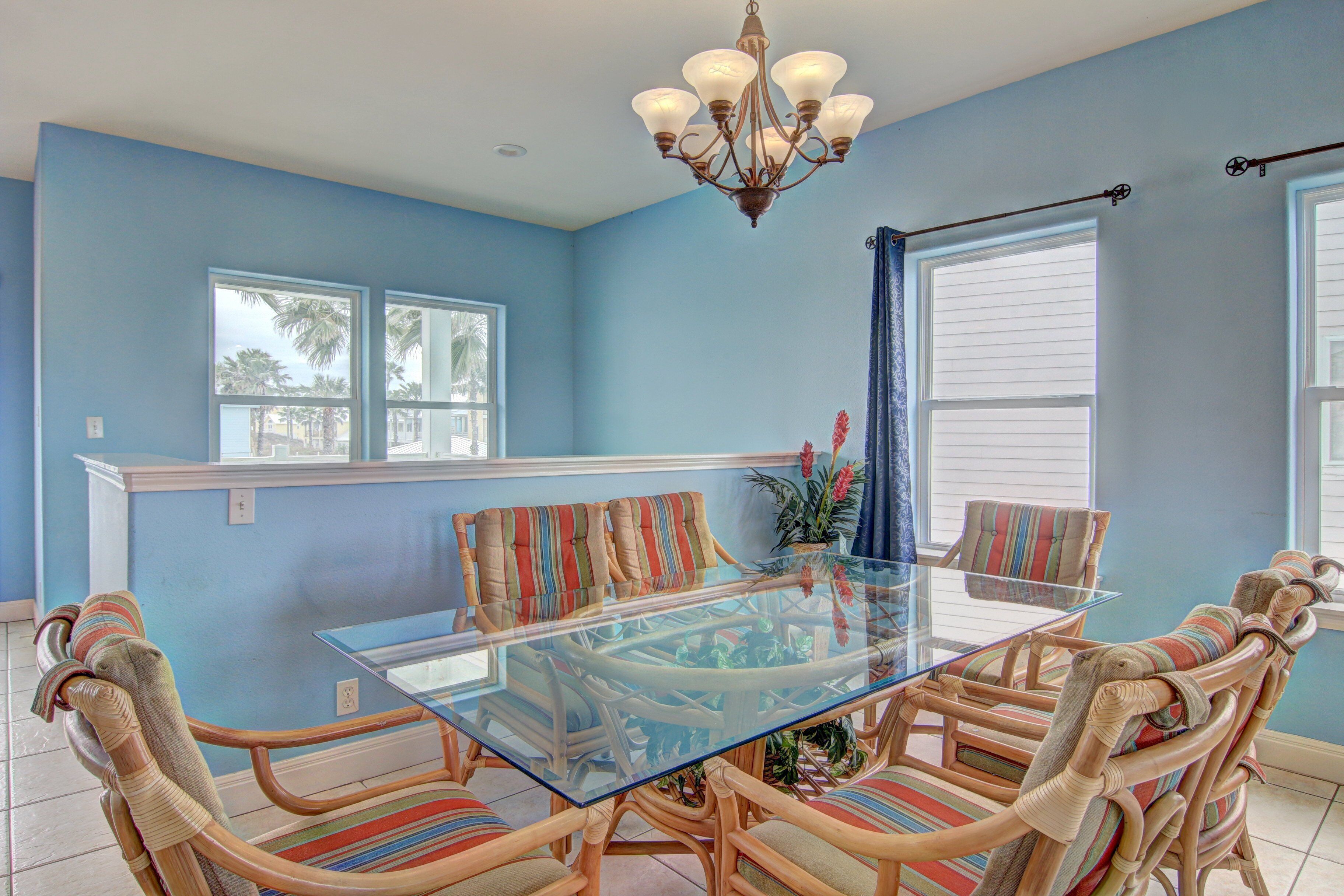 A dining area with a glass-top table surrounded by wicker chairs with striped cushions, a chandelier, and a window with blinds.
