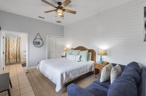 A bedroom with a bed, a blue couch, a wooden ceiling fan, and a nightstand with a lamp.