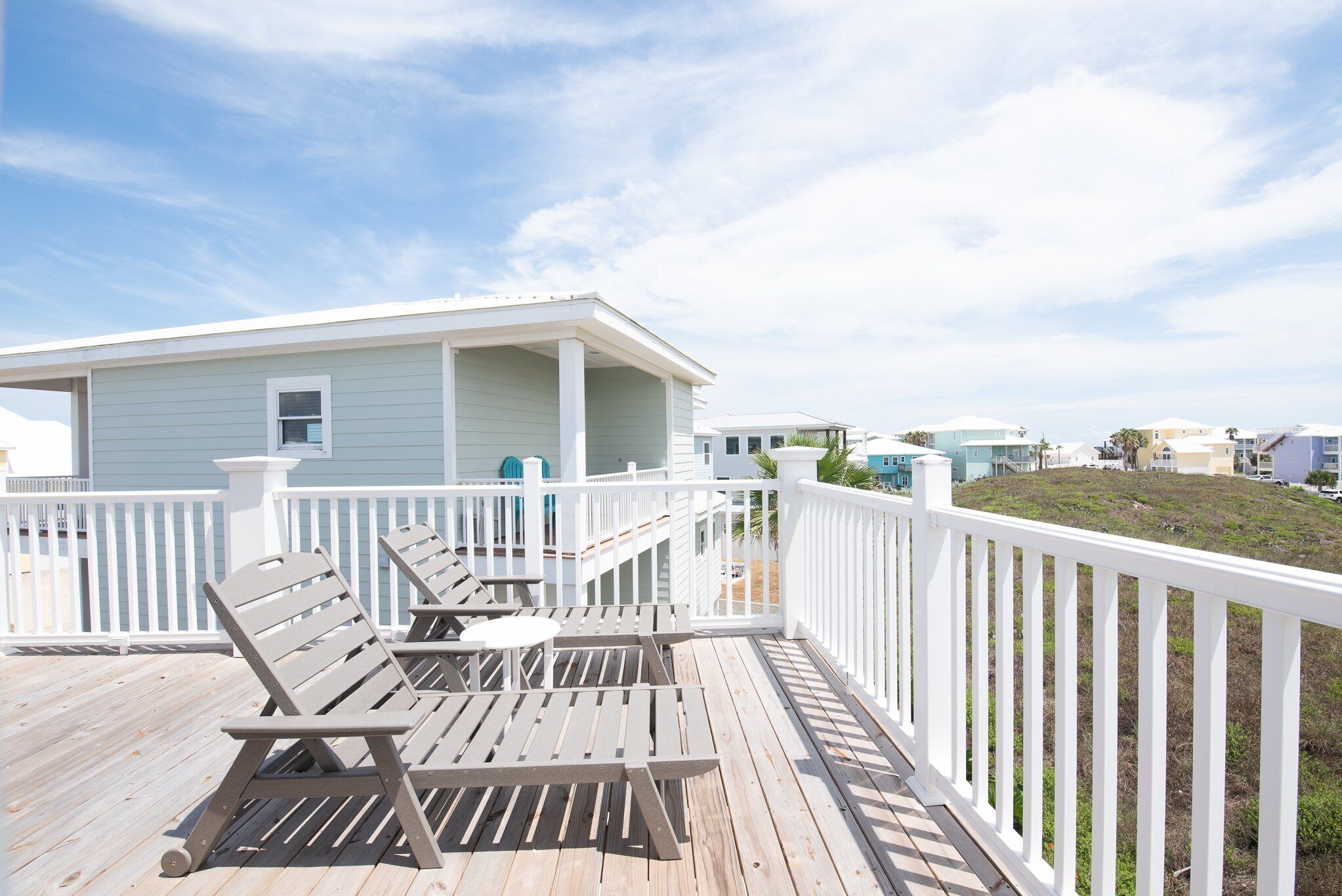 A wooden deck with two lounge chairs and a small table, a white railing, and a light blue building in the background.