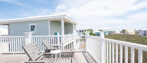 A wooden deck with two lounge chairs and a small table, a white railing, and a light blue building in the background.