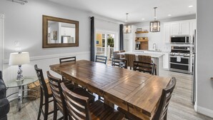 A dining area with a wooden table and chairs, a large mirror, and a view into a kitchen with white cabinets and a microwave. - Casa Azul - Boardwalk to the Beach! (Port Aransas)