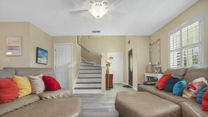 A living room with a beige couch, colorful pillows, a ceiling fan, and a staircase leading to another room. - Palma Verde - Pet Friendly! (Port Aransas)