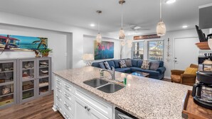 A modern kitchen with granite countertops, a double sink, and a dining area with a blue sofa and wicker chairs. - Sunrise Villas Condos (Port Aransas)