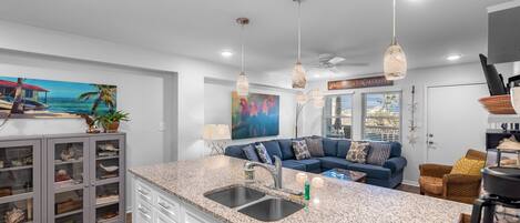 A modern kitchen with granite countertops, a double sink, and a dining area with a blue sofa and wicker chairs.