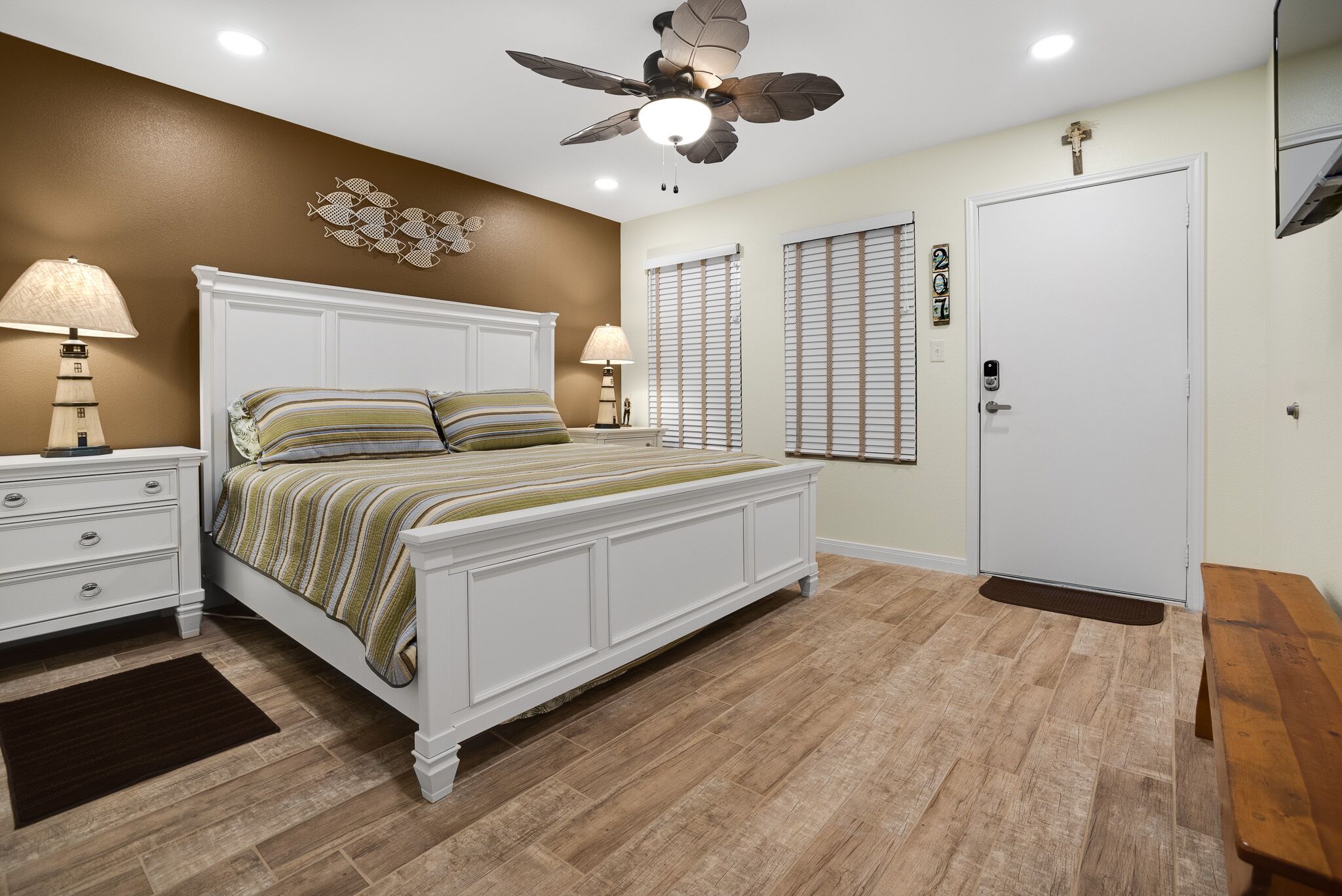 A bedroom with a white bed, two bedside tables with lamps, a ceiling fan, a wooden floor, and a door with a doormat.