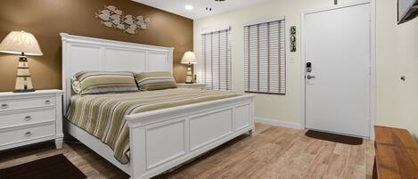 A bedroom with a white bed, two bedside tables with lamps, a ceiling fan, a wooden floor, and a door with a doormat.