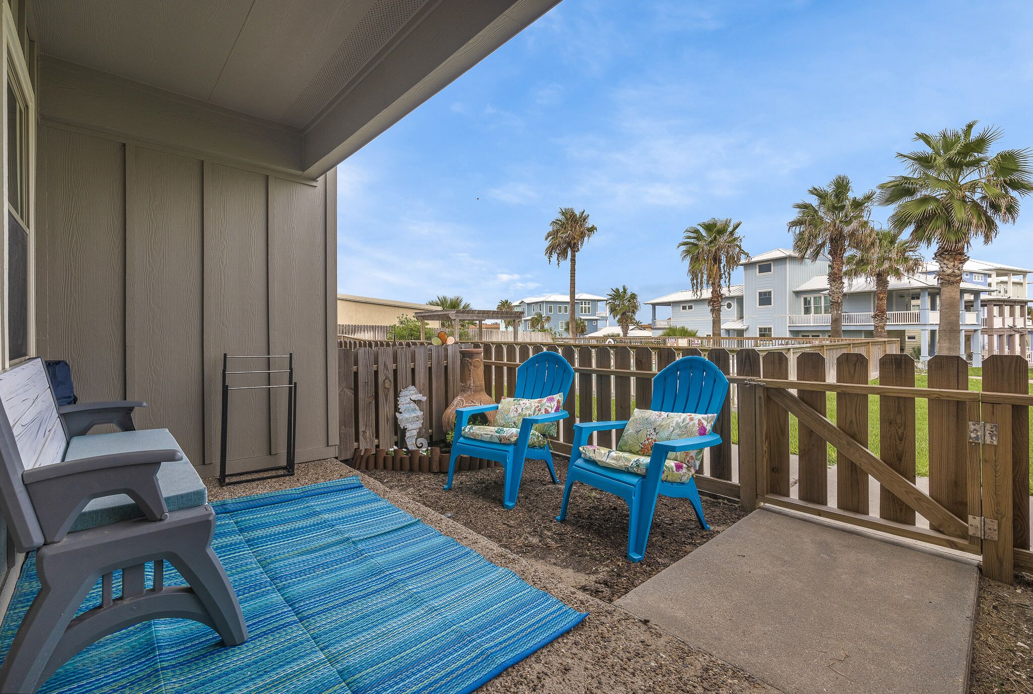 A covered patio with a blue mat, two blue Adirondack chairs, and a gray bench.