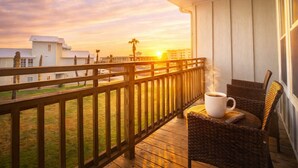 A balcony with wicker chairs, a white mug, and a view of buildings and palm trees during sunset. - Sunrise Villas Condos (Port Aransas)