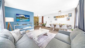 A modern living room with a large sofa, a coffee table, and a dining area with a table and chairs. There is a painting on the wall and a lamp on a side table. - Stocked Condo - Sleeps 10 - Palmwavestays (Coconut Creek)