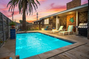 A swimming pool with lounge chairs and a barbecue grill, surrounded by a wooden deck and a brick building. - Oleander House - Pet Friendly! Private Pool! (Port Aransas)
