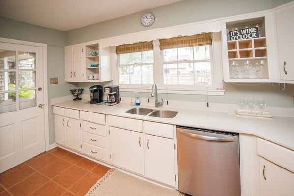 A kitchen with white cabinets, a dishwasher, a coffee maker, and a window with a wicker blind. - Oleander House - Pet Friendly! Private Pool! (Port Aransas)