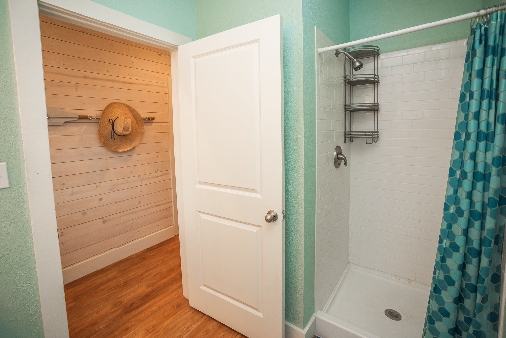 A bathroom with a shower, a wooden door, and a towel rack with a hat.