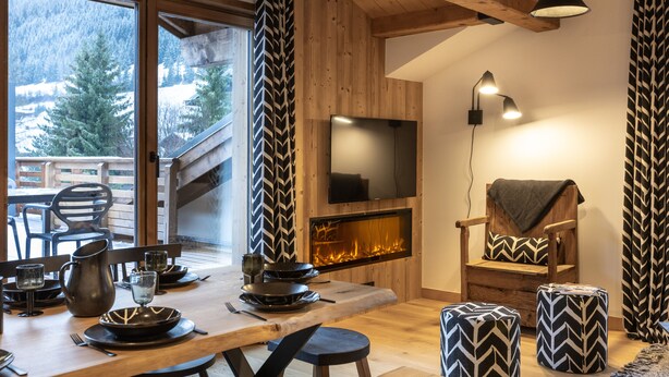 Un salon chaleureux avec une cheminée, des poutres en bois et une grande fenêtre donnant sur un paysage enneigé.