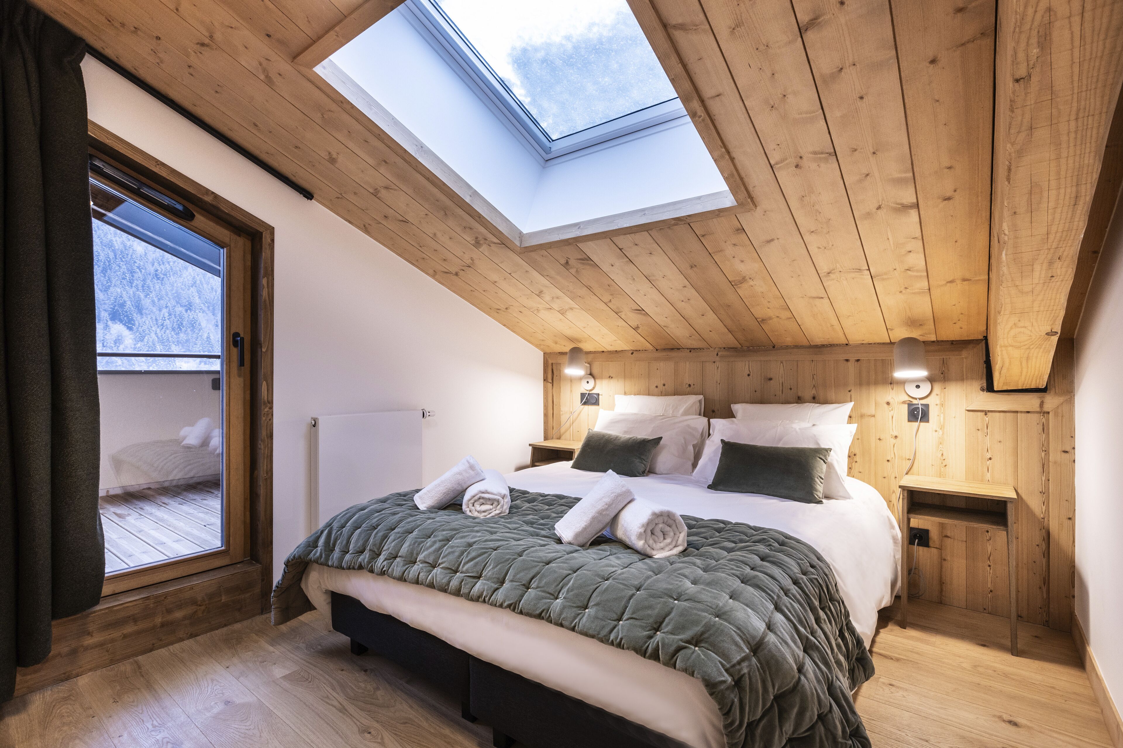 A bedroom with a skylight, wooden ceiling, a bed with a green quilt, two bedside tables with lamps, and a view through a window.