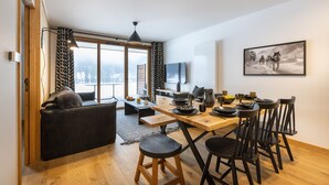 Un salon moderne avec un canapé, une table à manger et des chaises. On y trouve une grande fenêtre avec des rideaux, un téléviseur à écran plat et un tableau accroché au mur.