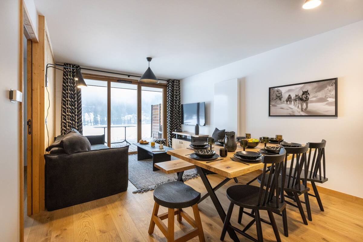 Ein modernes Wohnzimmer mit einer Couch, einem Esstisch und Stühlen. Ein großes Fenster mit Vorhängen, ein Flachbildfernseher und ein Gemälde an der Wand sind ebenfalls vorhanden.