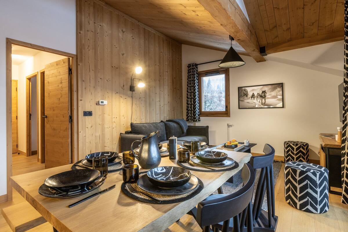 Ein Holzinterieur mit einem für zwei Personen gedecktem Esstisch, einer Couch und einem Fenster mit Blick auf Bäume.
