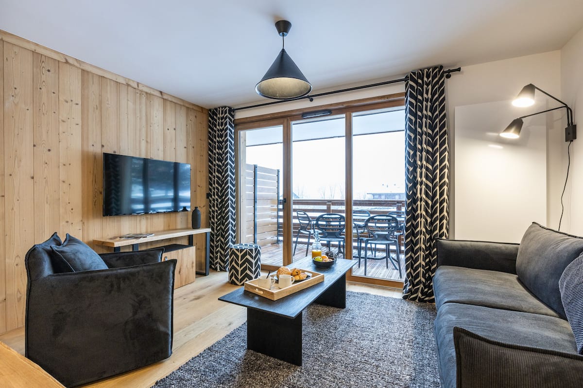 Ein modernes Wohnzimmer mit Flachbildfernseher, einer Couch, einem Couchtisch mit Frühstücksangeboten und einem großen Fenster mit Vorhängen.