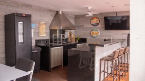A modern kitchen with a black refrigerator, stainless steel appliances, a large island with bar stools, and a flat-screen TV mounted on the wall. - Casa 5 Estrelas Top!!! All Incluse com 3 Funcionários (Praia de Maresias São Sebastião)