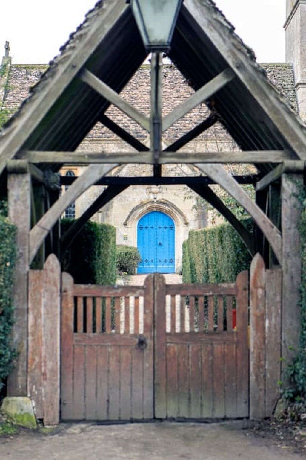 Une porte en bois surmontée d’une structure métallique formant un toit, qui donne accès à un bâtiment doté d’une porte bleue et de fenêtres en arc de cercle.