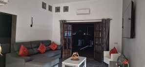 Sala de estar com um sofá cinza, uma mesa branca e um objeto decorativo vermelho. Há uma porta que dá para o exterior e uma televisão fixada na parede.