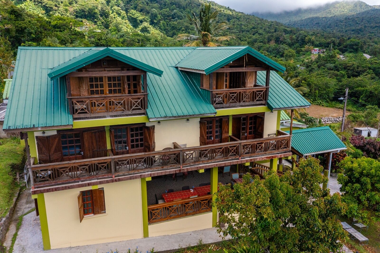 A two-story building with a green roof and wooden balconies, surrounded by lush greenery and hills.