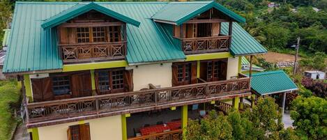A two-story building with a green roof and wooden balconies, surrounded by lush greenery and hills.