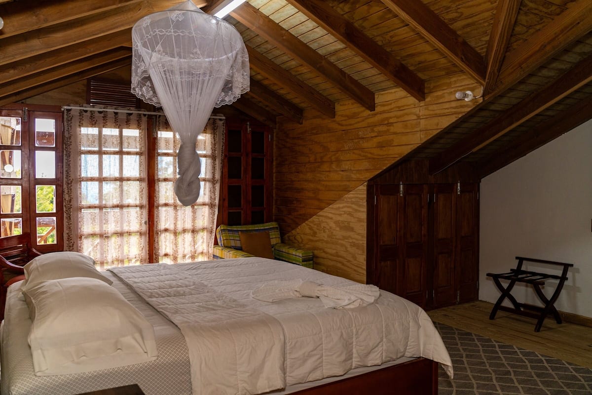A bedroom with a wooden ceiling, a bed with white bedding, a window with curtains, and a chair.