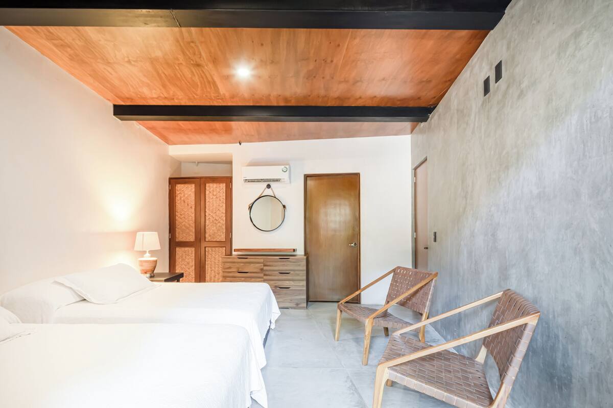 A hotel room with two beds, a wooden ceiling, a wall-mounted air conditioner, and a door leading to another room.