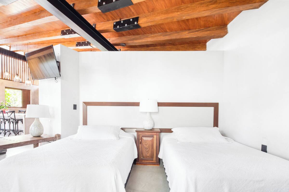 A bedroom with two beds, a wooden headboard, a bedside table with a lamp, and a window with blinds.
