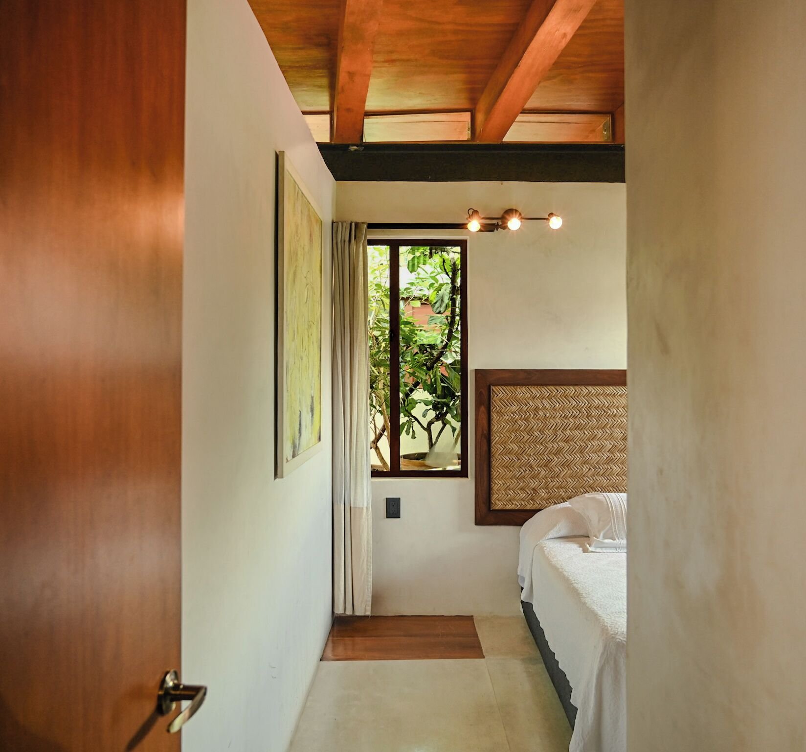 A narrow hallway with a wooden door, a bed with a woven headboard, a window with a view of greenery, and a light fixture above the bed.
