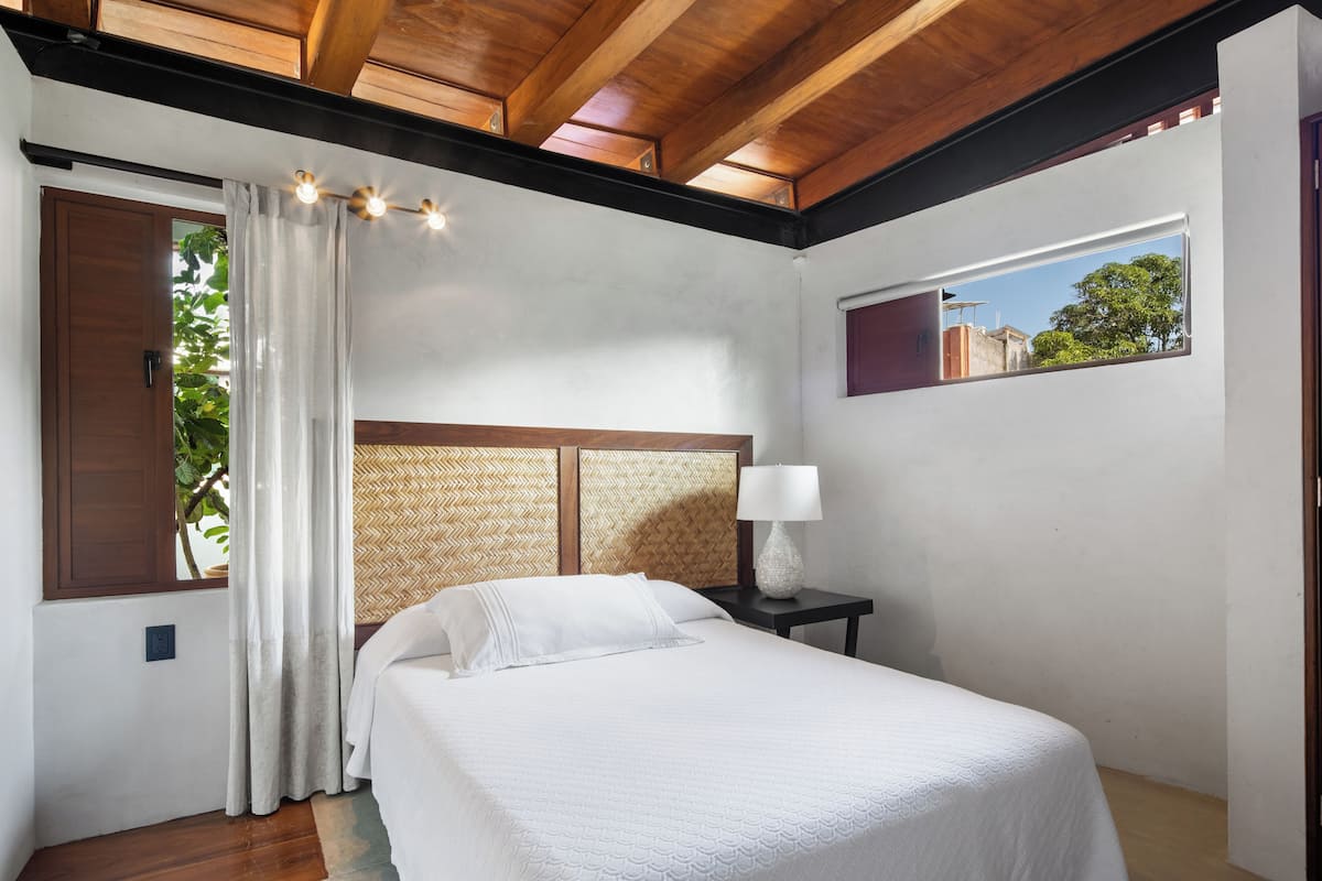 A bedroom with a bed, a nightstand, a lamp, a window, and a view of greenery outside.