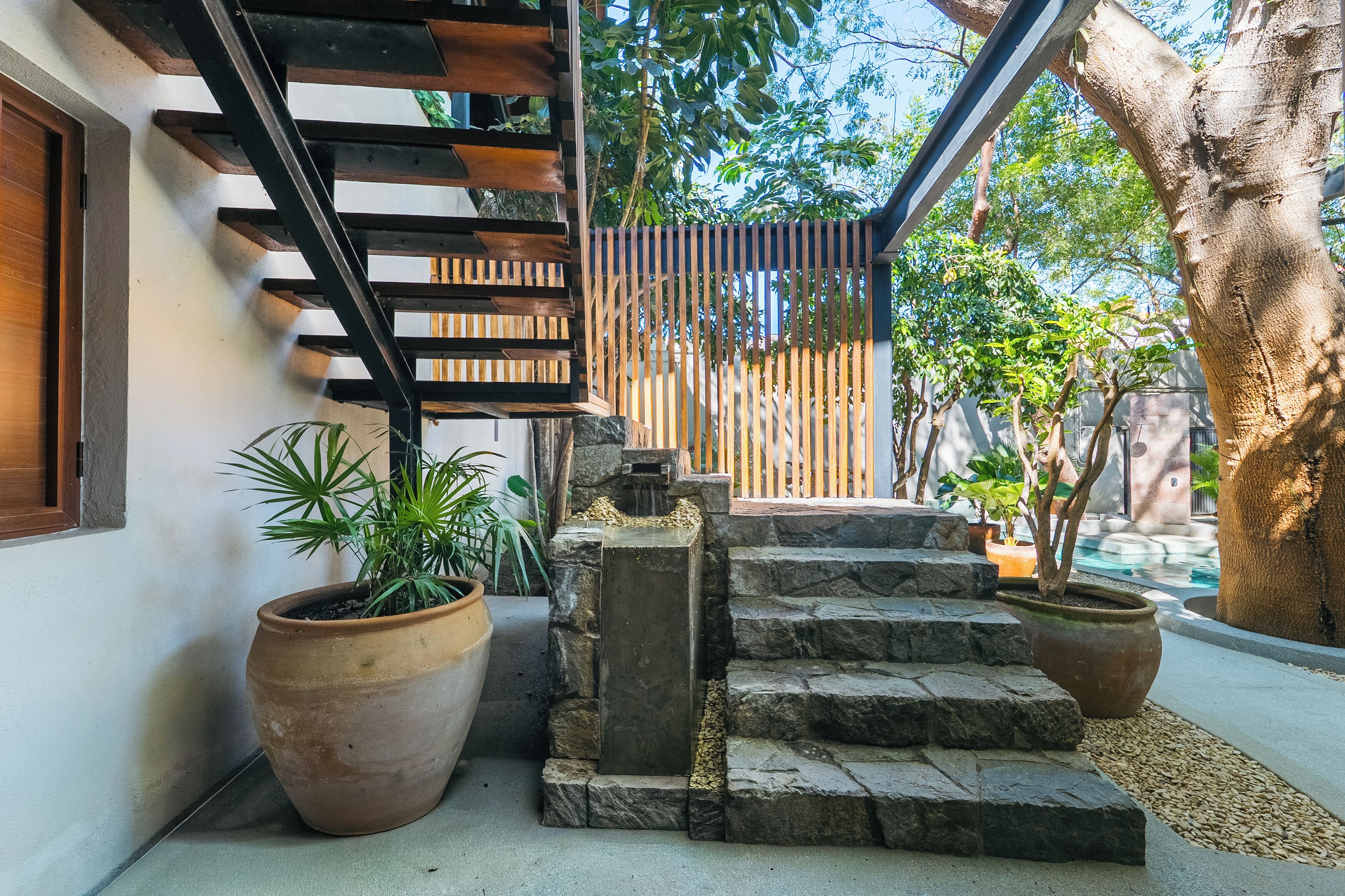 Una escalera de piedra conduce a una terraza de madera, rodeada de plantas en macetas y una valla metálica.