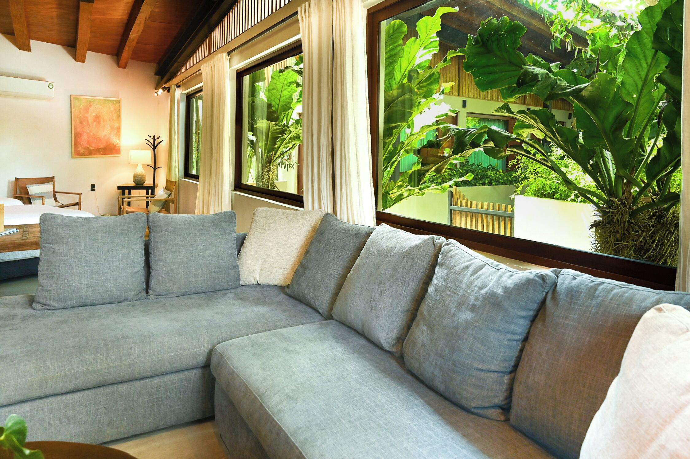 A modern living room with a grey sectional sofa, a wooden ceiling, and large windows with curtains.