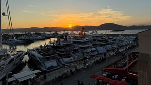 Ein Yachthafen mit vertäuten Booten, im Hintergrund ein Sonnenuntergang und ein Café mit roten Sonnenschirmen.