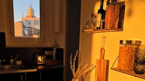 Eine Küche mit Fensterblick auf ein Gebäude mit einem Glockenturm, Holzregale mit Küchenutensilien und ein Holztisch mit Besteck.