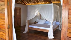 Un lit à baldaquin en bois, avec du linge de lit blanc et un dais, une porte en bois et une table de chevet avec une lampe.