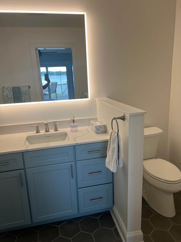 A bathroom with a white sink, blue cabinets, a toilet, and a large mirror with a view of a balcony.