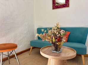 A teal sofa, a round wooden table with a vase of flowers, and a chair with metal legs.