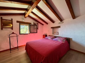 A bedroom with a bed, wooden beams, a window, and a wall-mounted shelf.