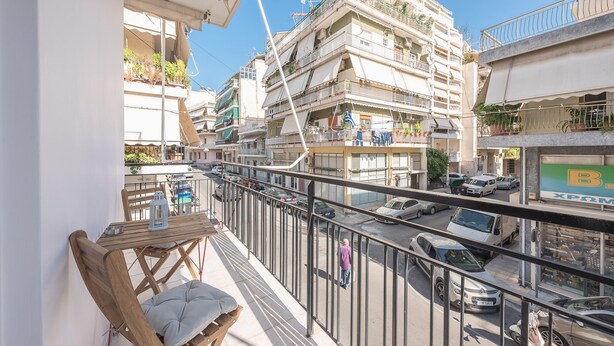 Un balcon avec une table en bois et un banc rembourré, donnant sur une rue animée, bordée de voitures et d’immeubles.