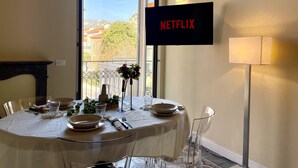A dining area with a table set for four, a TV screen displaying 'NETFLIX', and a view of buildings and trees through a large window. - [villa Migliorini] Lusso & Comfort in Finalborgo (Finale Ligure)