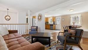 Uma sala de estar ampla com sofá, poltrona e mesa de centro, que dá acesso a uma cozinha e área de jantar.